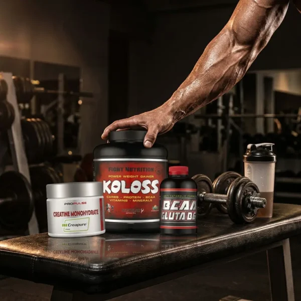 Homme musclé dans une salle de sport posant une sélection de compléments alimentaires sur un banc: gainer Power Weight KOLOSS, créatine monohydrate, et autres flacons, près d’un haltère. Alt texte inclut:PACK MASSE RAPIDE.
