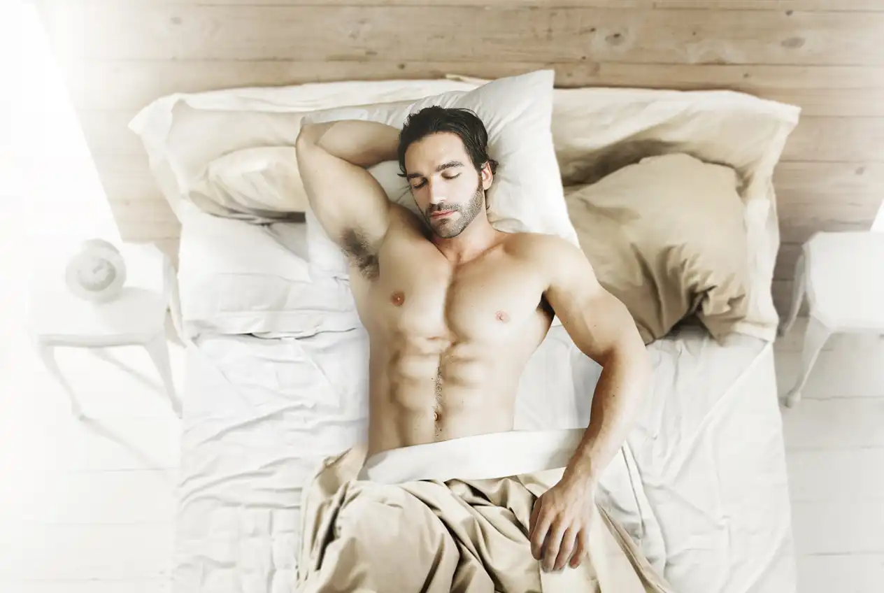 Homme musclé dormant sur lit, torses et abdominaux visibles, couvert partiellement par une couverture beige.